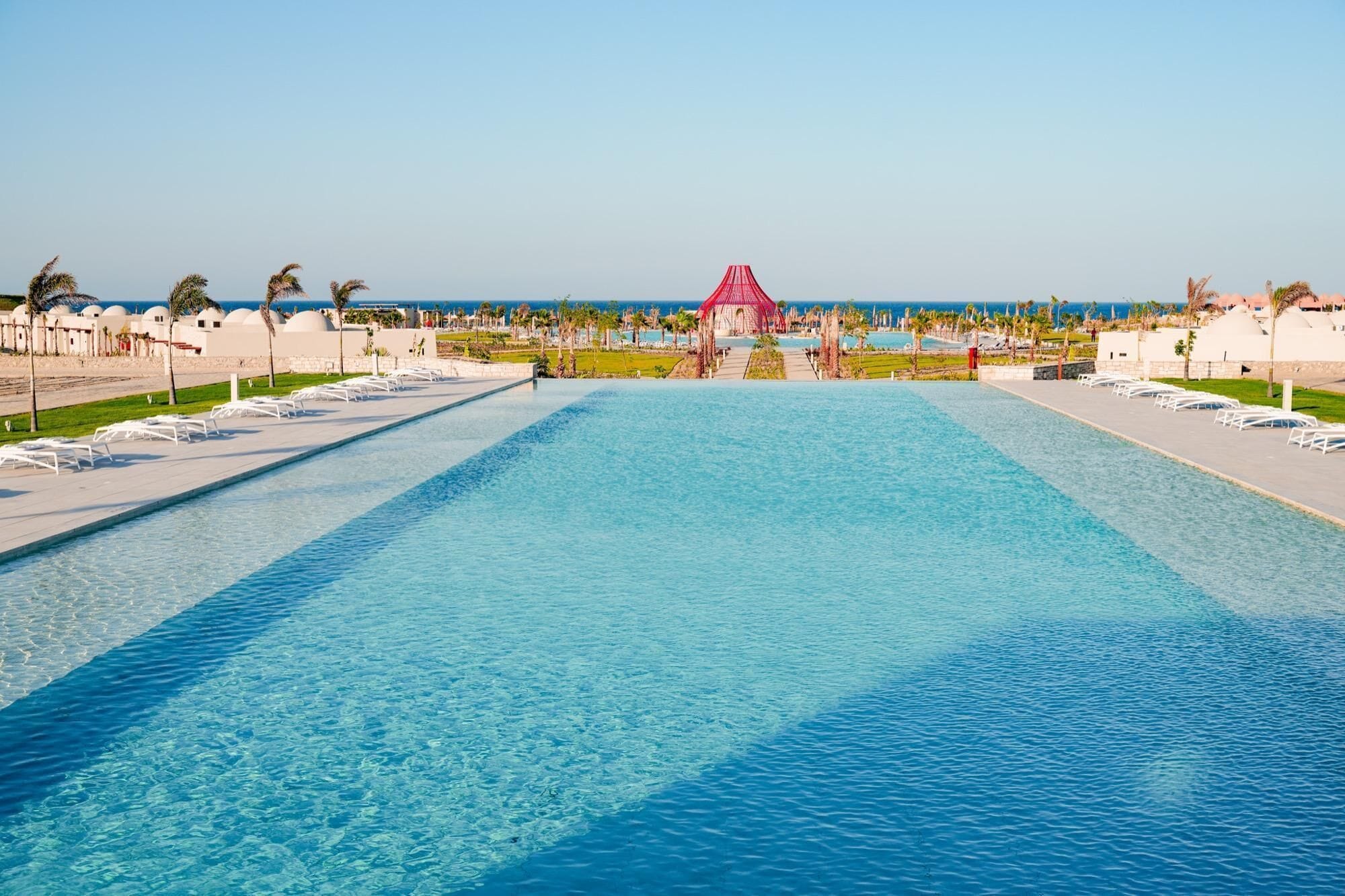 A large rectangular pool with a clear blue water surface, surrounded by white lounge chairs and palm trees.