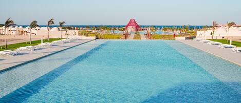 A large rectangular pool with a clear blue water surface, surrounded by white lounge chairs and palm trees.