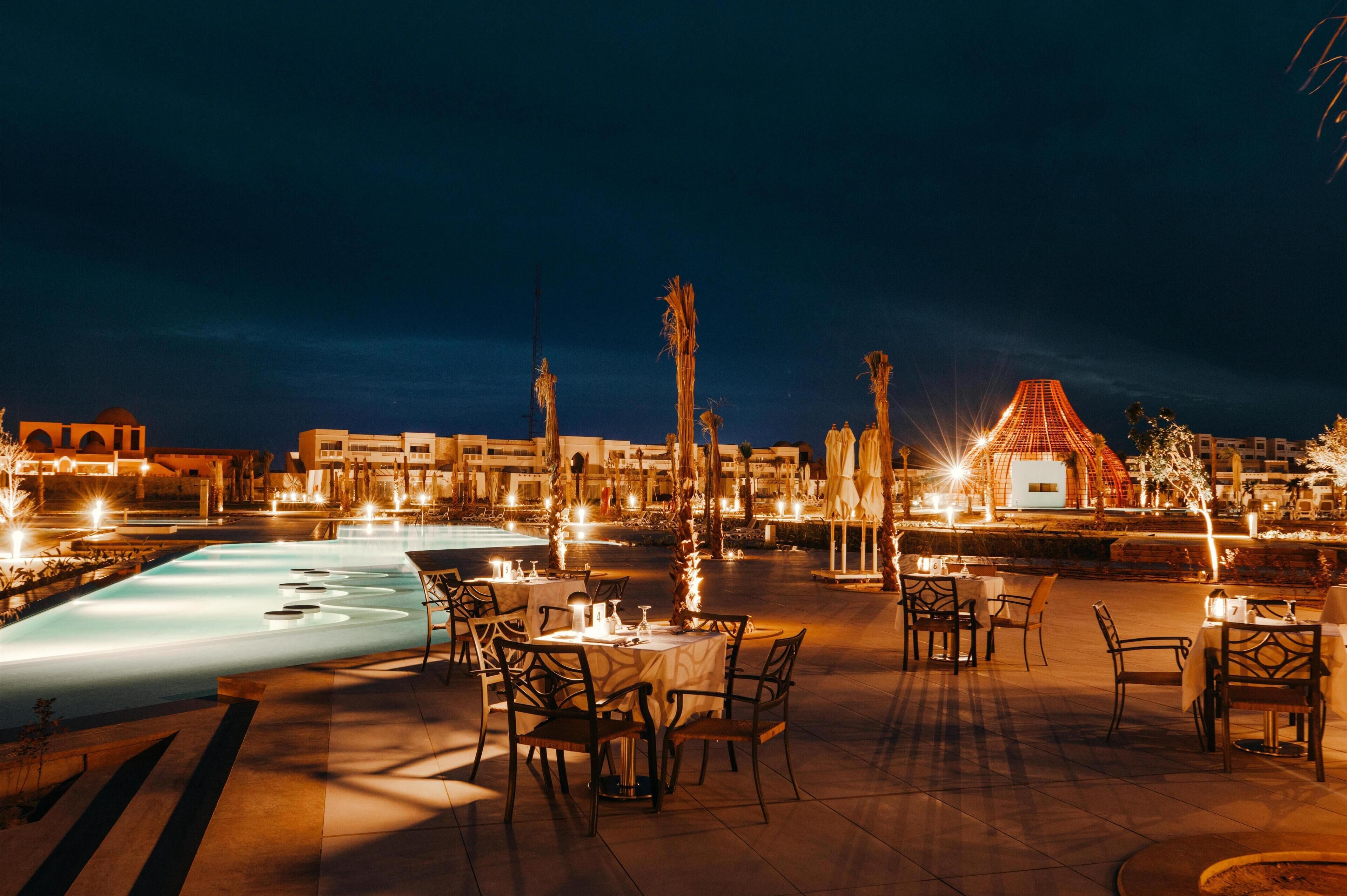 Un espace extérieur aménagé pour les repas, avec des tables et des chaises, une piscine et, au fond, un bâtiment doté d’un toit remarquable.