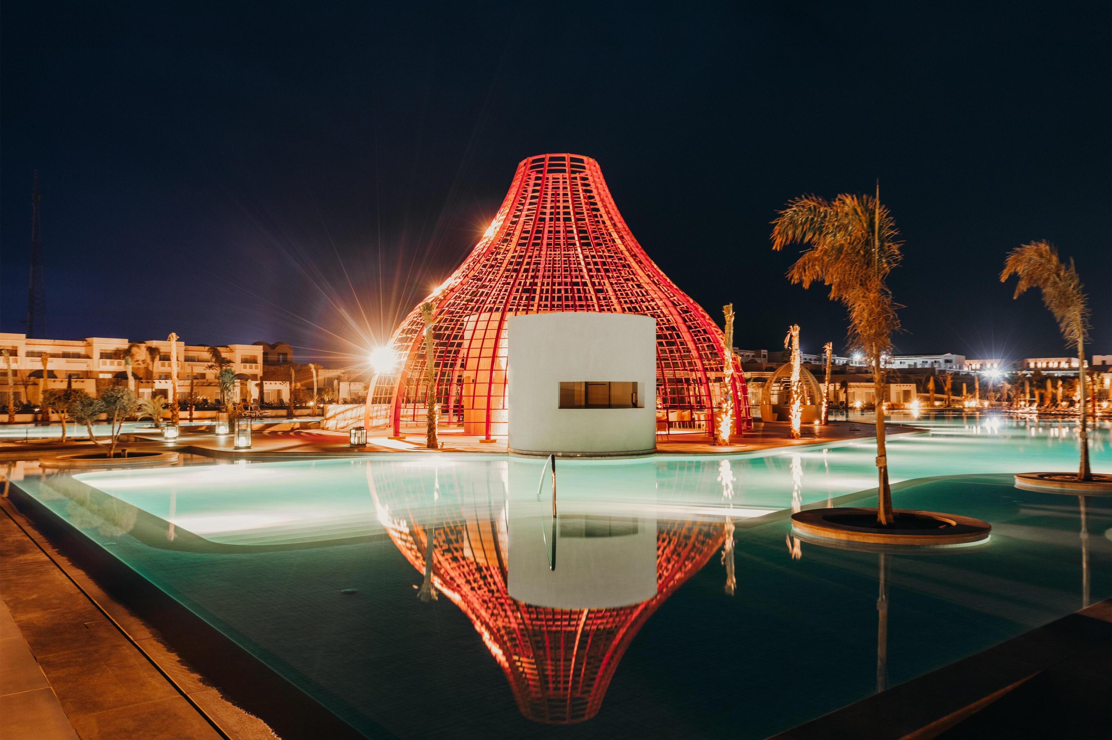 A modern hotel with a unique red-lit structure, a swimming pool, and palm trees.