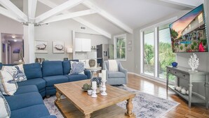 Un salón amplio con techo alto, un ventanal con persianas, un sofá azul, una mesa de centro de madera y un televisor sobre un mueble.