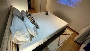 Une chambre à coucher avec un lit, un écran de projection diffusant une scène de plage, et un couloir menant à une autre pièce.