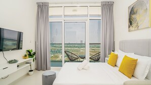 A modern hotel room with a large bed, a TV, a balcony with a city view, and a dining area with chairs. - Tanin - Sleek Studio Offering Unobstructed City Views (Dubai)