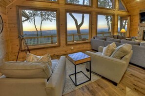 A cozy cabin interior with a sofa, armchair, and a small table. Large windows offer views of trees and a distant landscape.