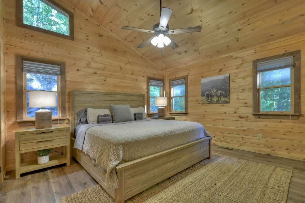 A wooden cabin bedroom with a bed, nightstands, a ceiling fan, and windows.