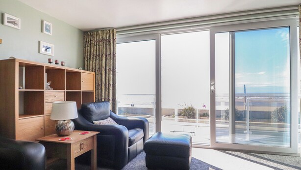 En moderne stue med sofa, skammel og et træskab. En stor skydedør fører ud til en balkon med udsigt over stranden.