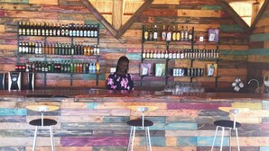 Un coin bar avec un mur en bois, plusieurs étagères remplies de bouteilles et trois tabourets.