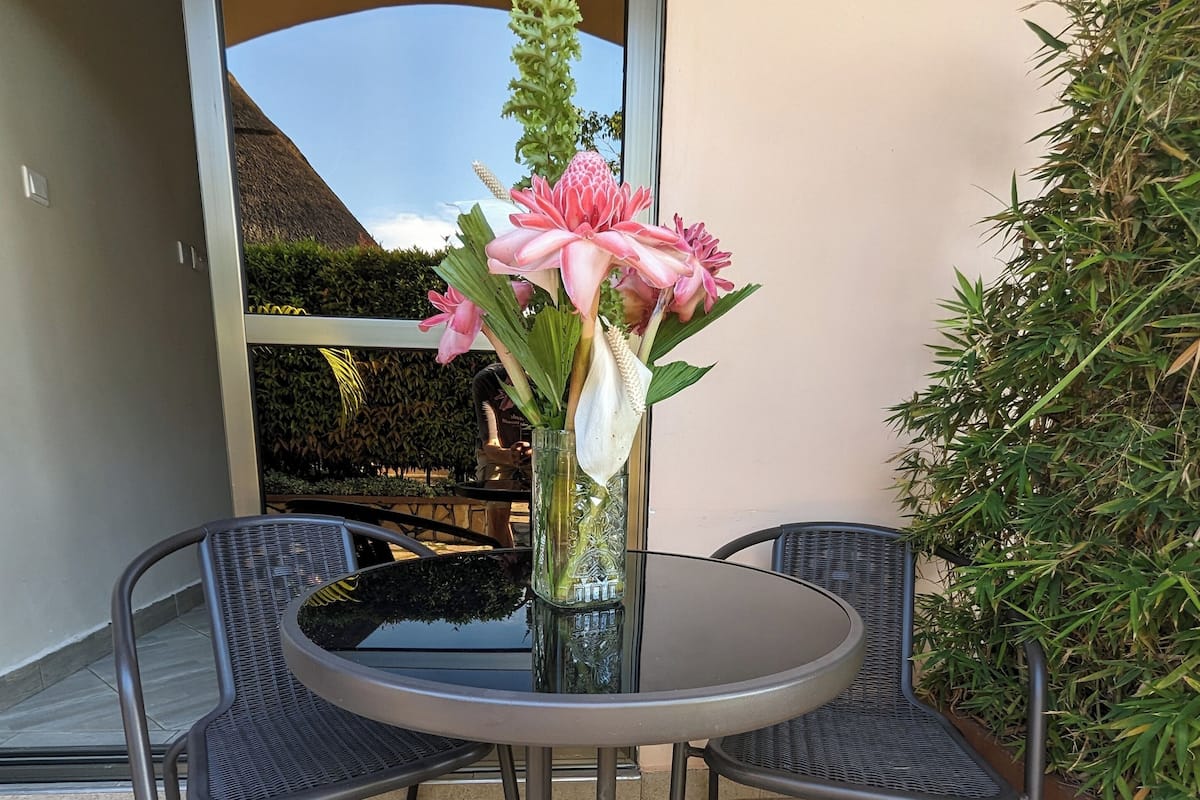 A round metal table with a glass vase containing pink flowers, surrounded by two metal chairs.