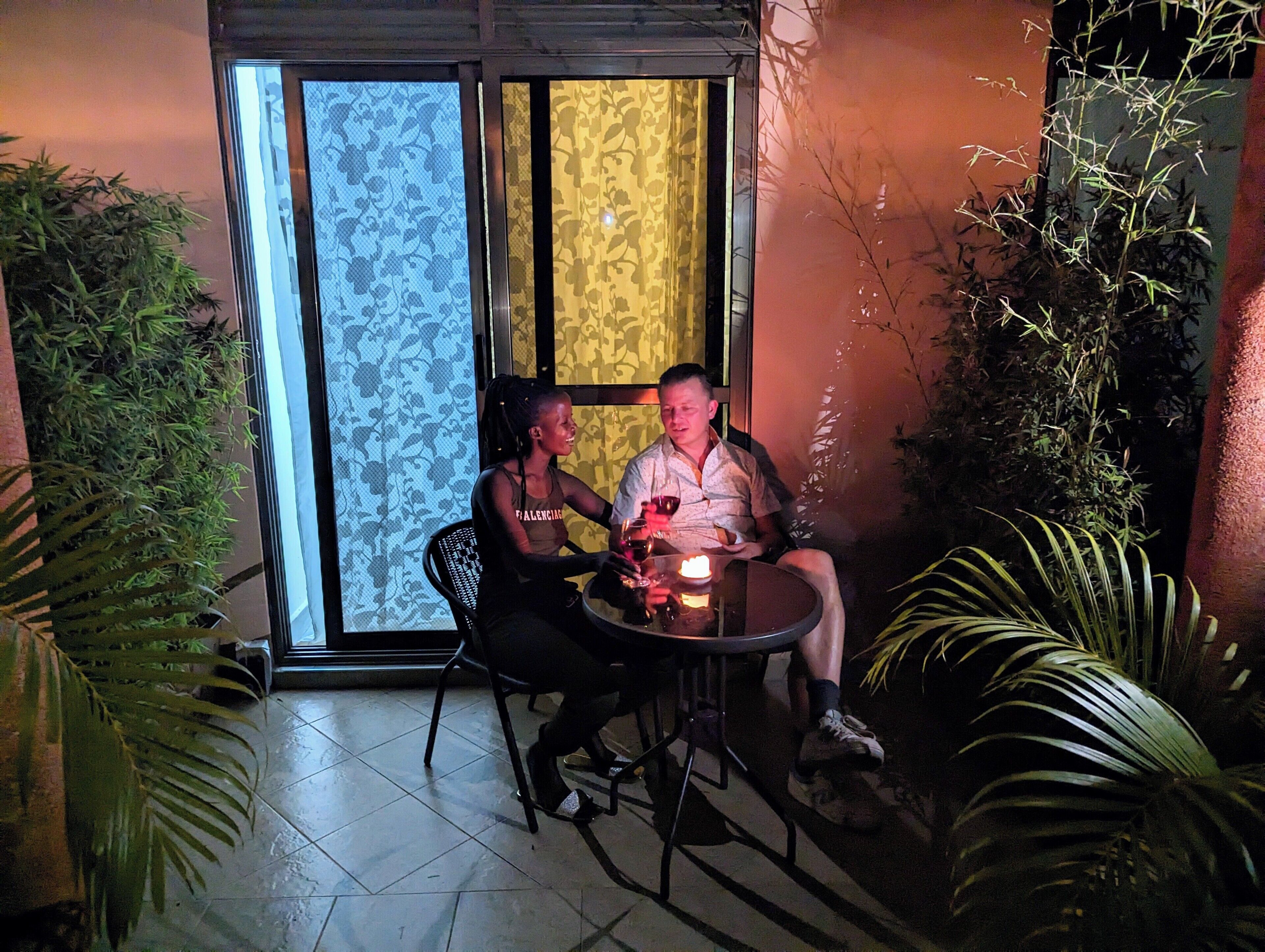 Two people seated at a table with a candle, surrounded by plants, in a room with patterned curtains and a tiled floor.