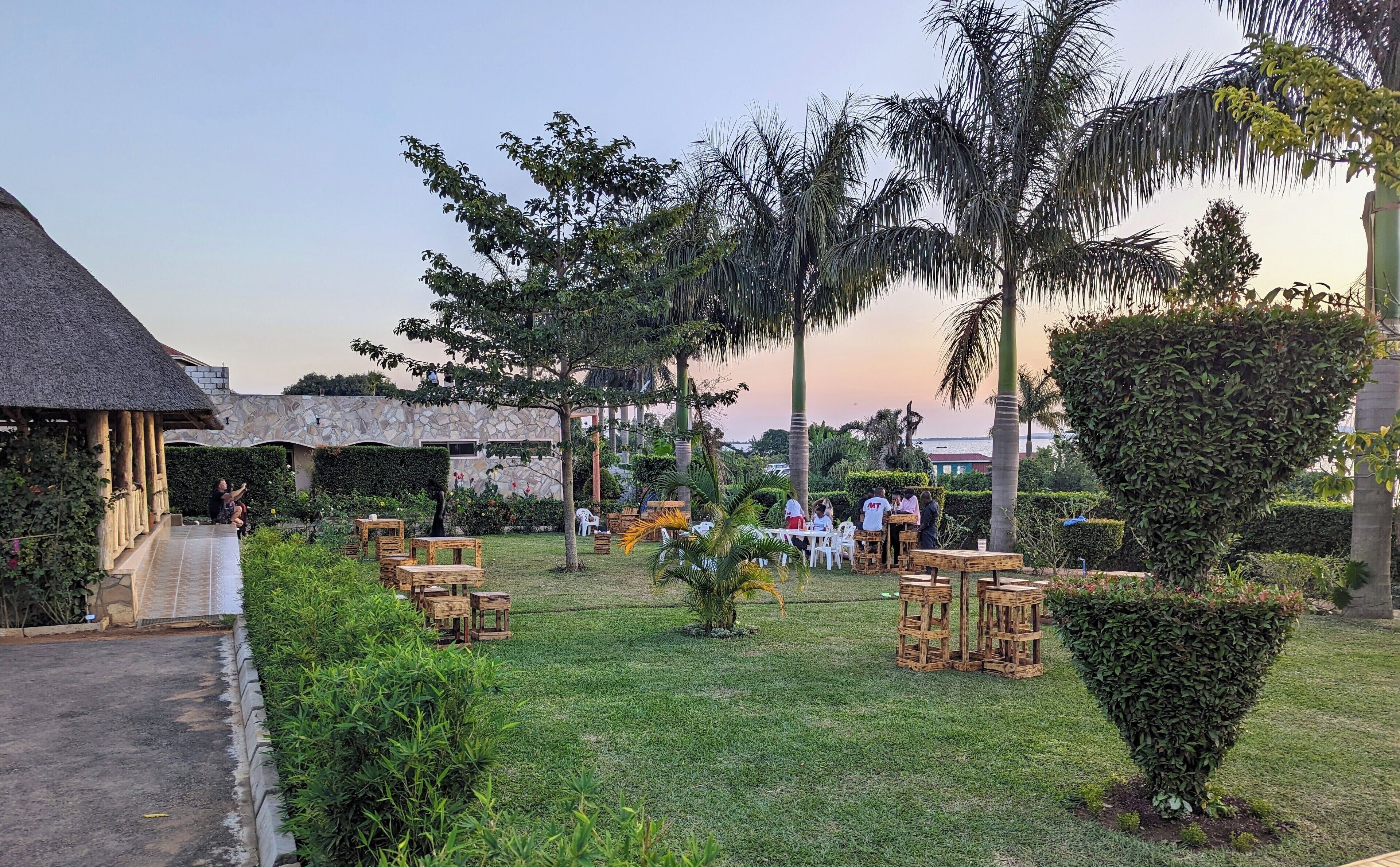 An outdoor dining area with wooden tables and chairs, surrounded by greenery and palm trees.
