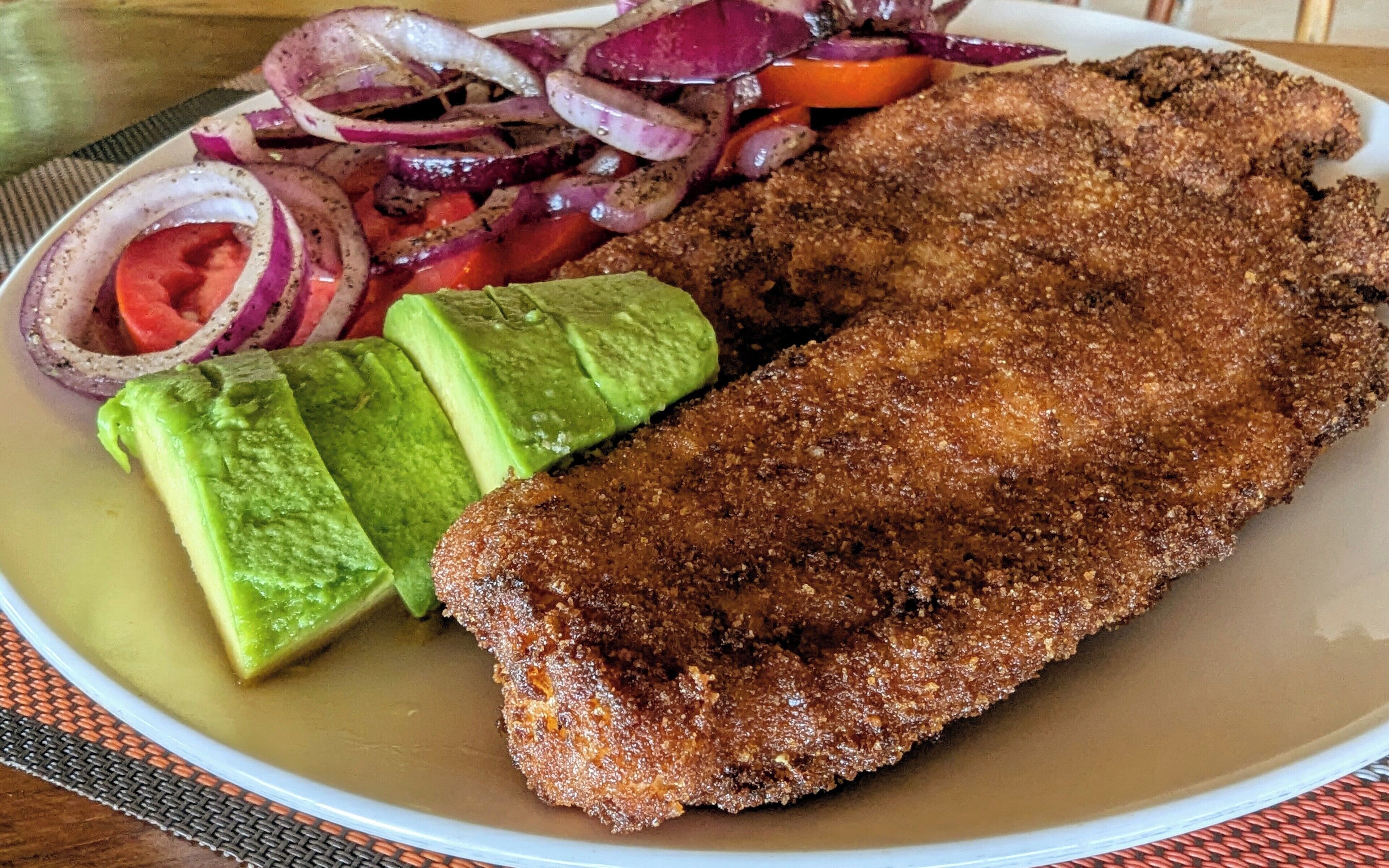 A plate with a breaded and fried item, avocado slices, and a side of sliced onions and tomatoes.