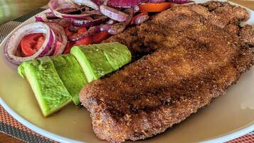 A plate with a breaded and fried item, avocado slices, and a side of sliced onions and tomatoes.