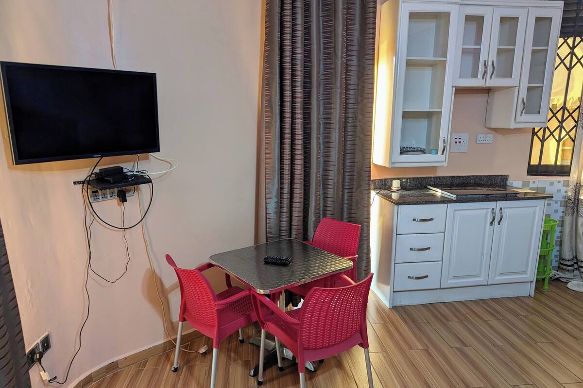 A small kitchenette with white cabinets and a countertop, a dining area with a table and red chairs, and a flat-screen TV mounted on the wall.