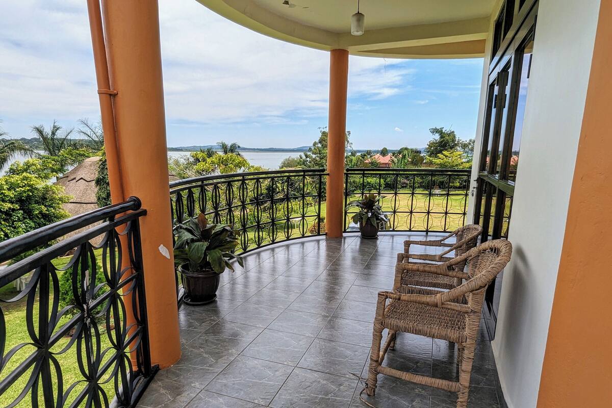 A covered balcony with two wicker chairs, a potted plant, and a view of a green landscape and distant hills.