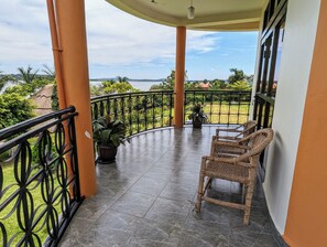 Un balcón techado con dos sillones de mimbre, una planta en maceta y vistas a un paisaje verde y colinas lejanas.