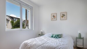 A bedroom with a large window, a bed with a white duvet, two bedside tables with lamps, and two framed botanical prints on the wall. - Modern 8-guest house with garden & BBQ (La Baule-Escoublac)