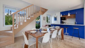 A modern kitchen with a wooden staircase, a dining area with white chairs, and a blue accent wall. - Modern 8-guest house with garden & BBQ (La Baule-Escoublac)