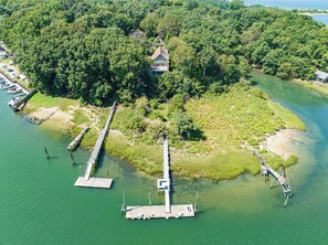 An aerial view of a secluded house surrounded by dense forest, connected to the mainland by a wooden dock.