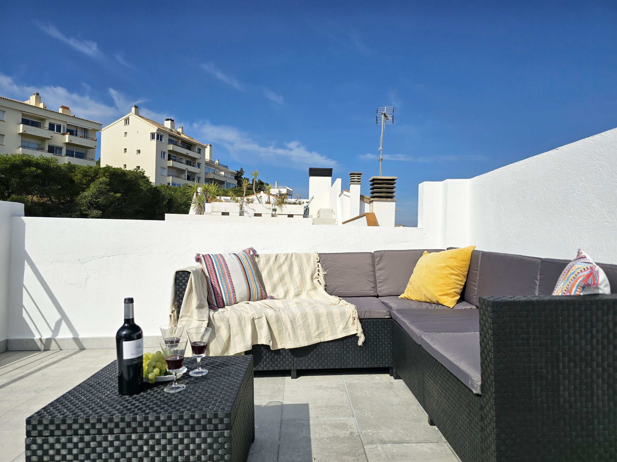 A rooftop terrace with a sofa set, a table with wine glasses and a bottle, and a view of buildings in the background.