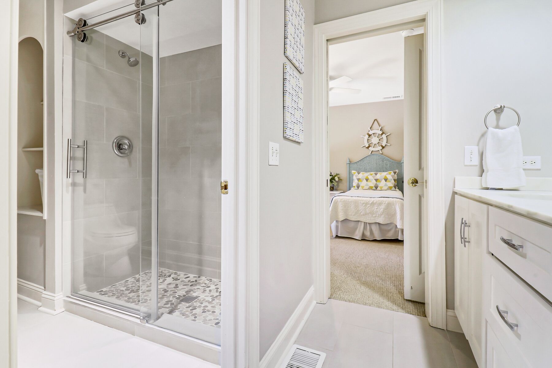 A modern bathroom with a glass-enclosed shower, a white vanity with a mirror, and a bed in the adjacent room.