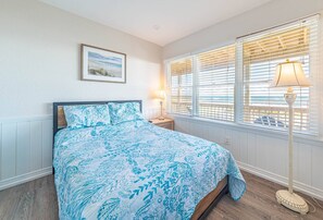 Een slaapkamer met een bed, een nachtkastje, een lamp, een raam met zonwering en een lijst met een afbeelding aan de muur.