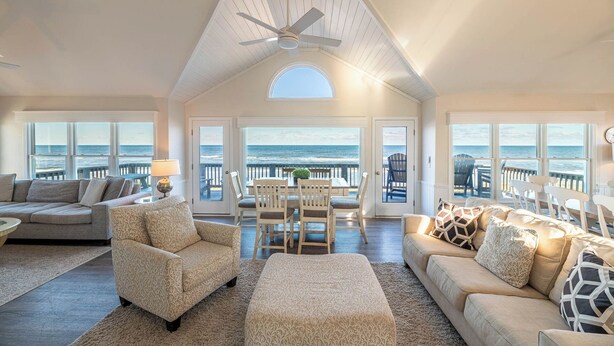 A spacious living room with a high ceiling, large windows offering ocean views, a ceiling fan, and a cozy seating area with a sofa and chairs.