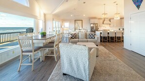 A spacious living area with a large window overlooking the ocean, a dining area with a wooden table and chairs, and a kitchen with modern appliances and pendant lighting.