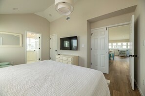 A bedroom with a large bed, a TV mounted on the wall, and a door leading to a kitchen area. - Sunrise Dream - Nags Head MP 16, Oceanfront, Ocean View, Pool, Hot Tub, Sauna, Pets OK, Flex Stays (Nags Head)