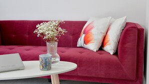 Un sofá rojo con dos almohadas, un florero transparente con flores blancas, una taza estampada y una computadora portátil sobre una mesa blanca.