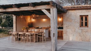 Eine hölzerne Terrasse im Freien mit einem rustikalen Steinhaus, einem Holztisch mit Stühlen und hängenden Lampen.