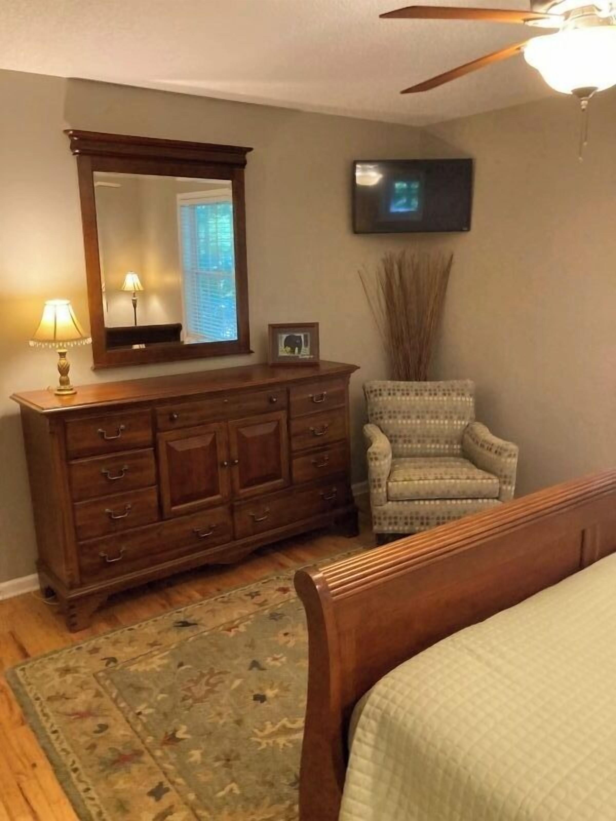 A bedroom with a wooden dresser, a chair, a bed, a ceiling fan, and a television mounted on the wall.