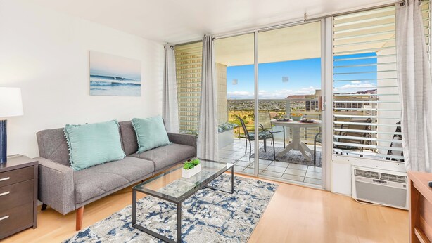 A modern living room with a grey sofa, a glass coffee table, and a view of a cityscape through large sliding glass doors. - Ocean-Golf Course Couples Retreat at Island Colony Short Walk to the Beach (Honolulu)