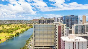 A high-rise building with multiple floors and balconies, situated next to a river with a park and residential area in the background. - Ocean-Golf Course Couples Retreat at Island Colony Short Walk to the Beach (Honolulu)