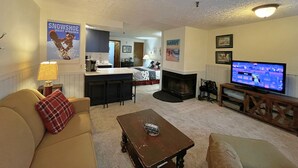 A living room with a sofa, coffee table, TV, fireplace, kitchen area, and a poster on the wall. - Ridgewood 54 - Snowshoe, Short walk to the Powderidge Lift! (Snowshoe)