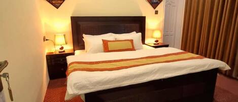 A hotel room with a large bed, two bedside tables with lamps, a door with a keyhole, and a curtain on the window.