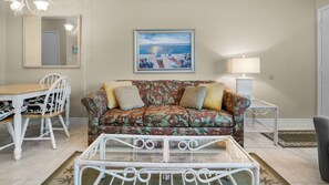 Ein Wohnzimmer mit einem blumigen Sofa, einem Glastisch, einem Esstisch mit Stühlen, einem Gemälde einer Strandansicht und einer Lampe.