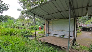 A covered outdoor seating area with a wooden bench, surrounded by lush greenery and a metal roof. - Hotel O Jati Homestay Near Taman Suranadi (Selong Belanak)