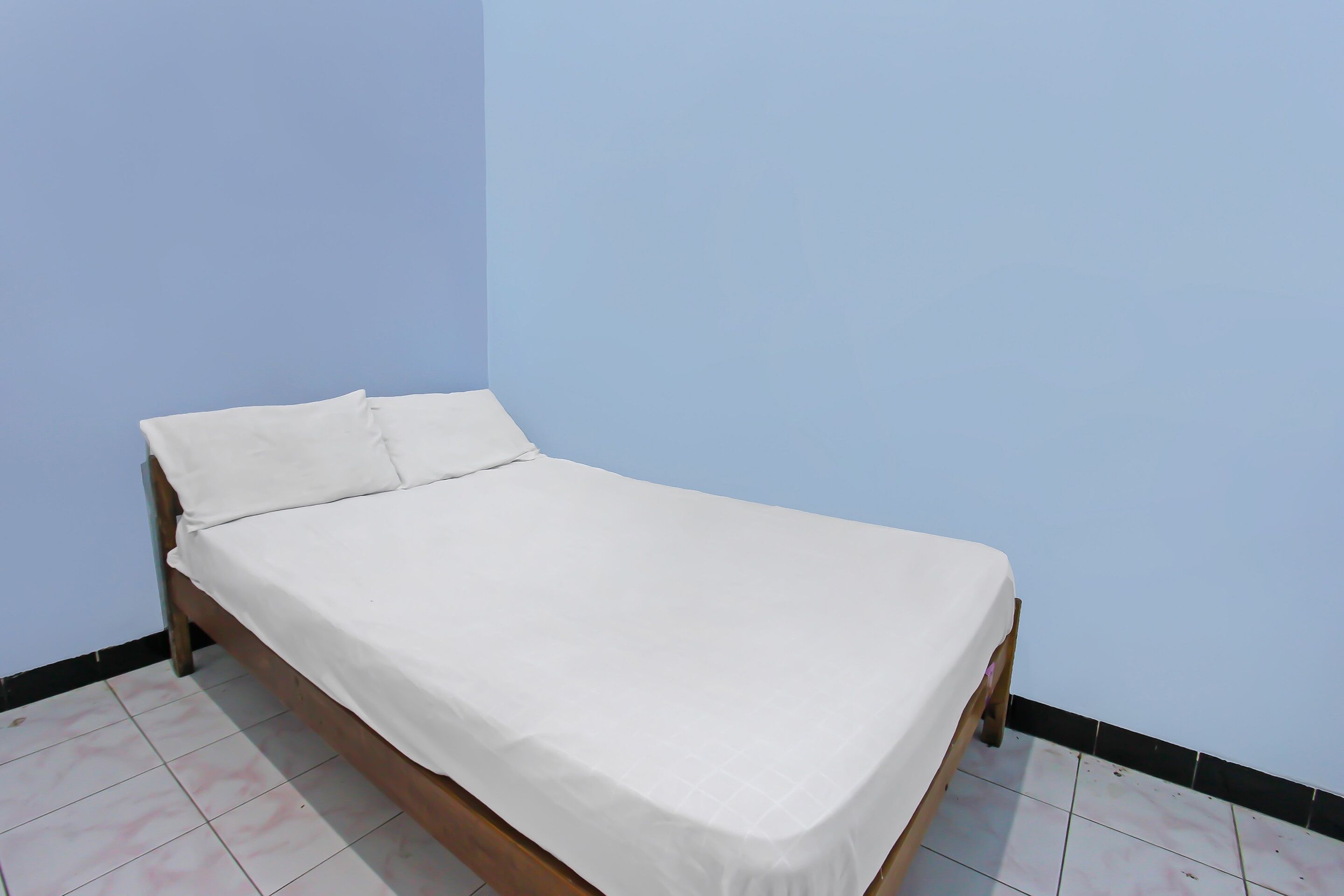Uma cama de solteiro com colchão e almofadas brancas num quarto com paredes azul claro e chão em mosaico.