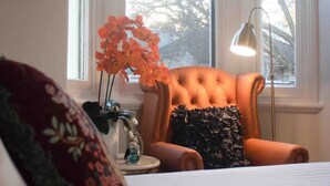 Un dormitorio con un ventanal grande, un sillón capitonado, una mesita de noche con una lámpara y un edredón estampado con motivos florales.