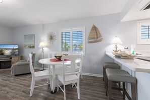 Un coin repas avec une table et des chaises blanches, un téléviseur et une fenêtre avec des stores.