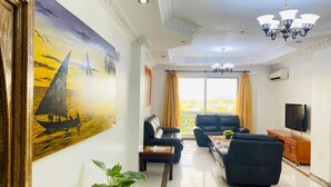 A living room with a large painting, a chandelier, a television, and a view of the outdoors. - Near the ocean  with Swimming pool ,Supermarket and ATM machine at the premises. (Dar es Salaam)