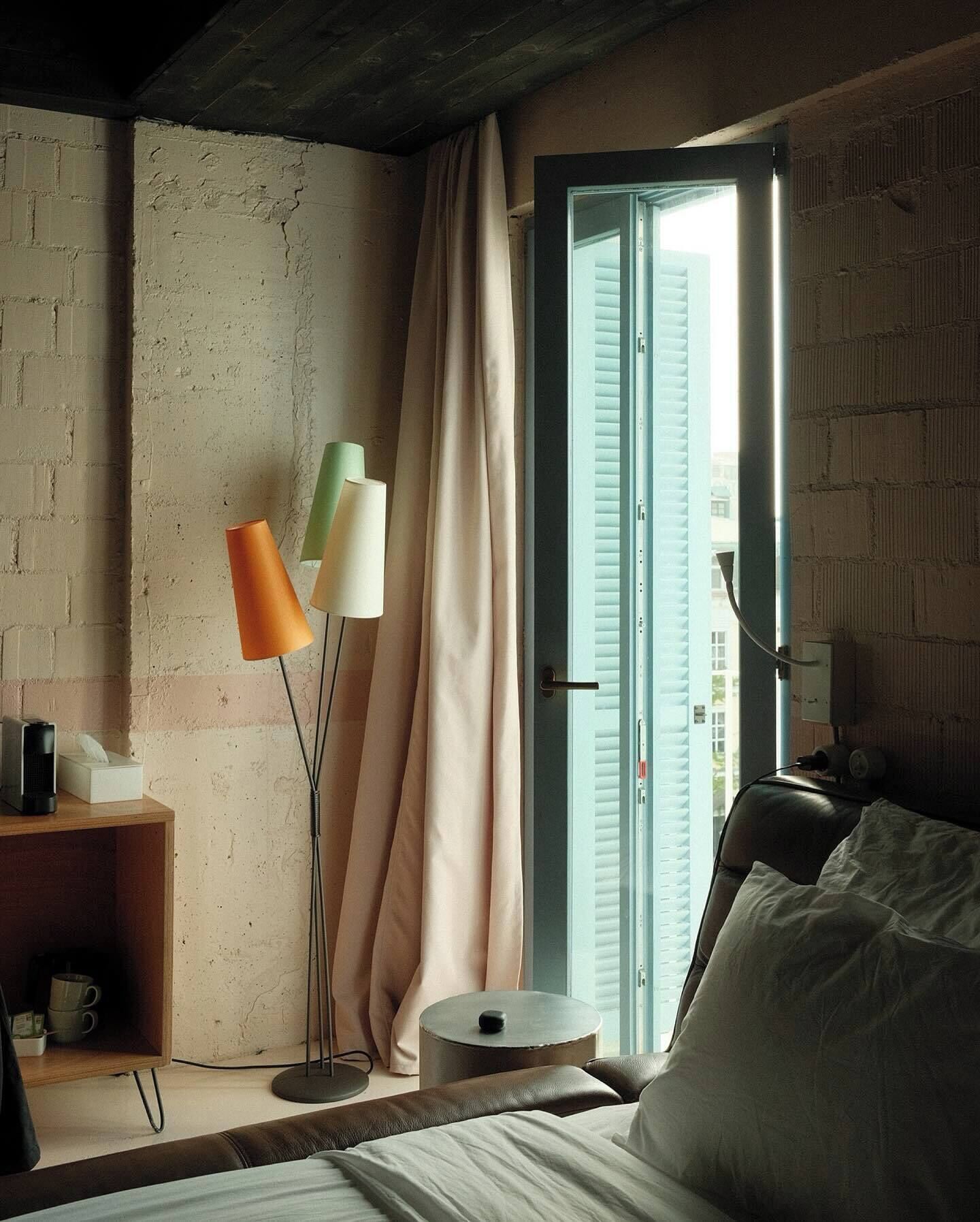Una camera da letto con un letto, un comodino con una lampada, una finestra con tende e vista sugli edifici esterni.