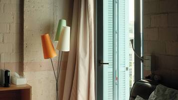 Una camera da letto con un letto, un comodino con una lampada, una finestra con tende e vista sugli edifici esterni.