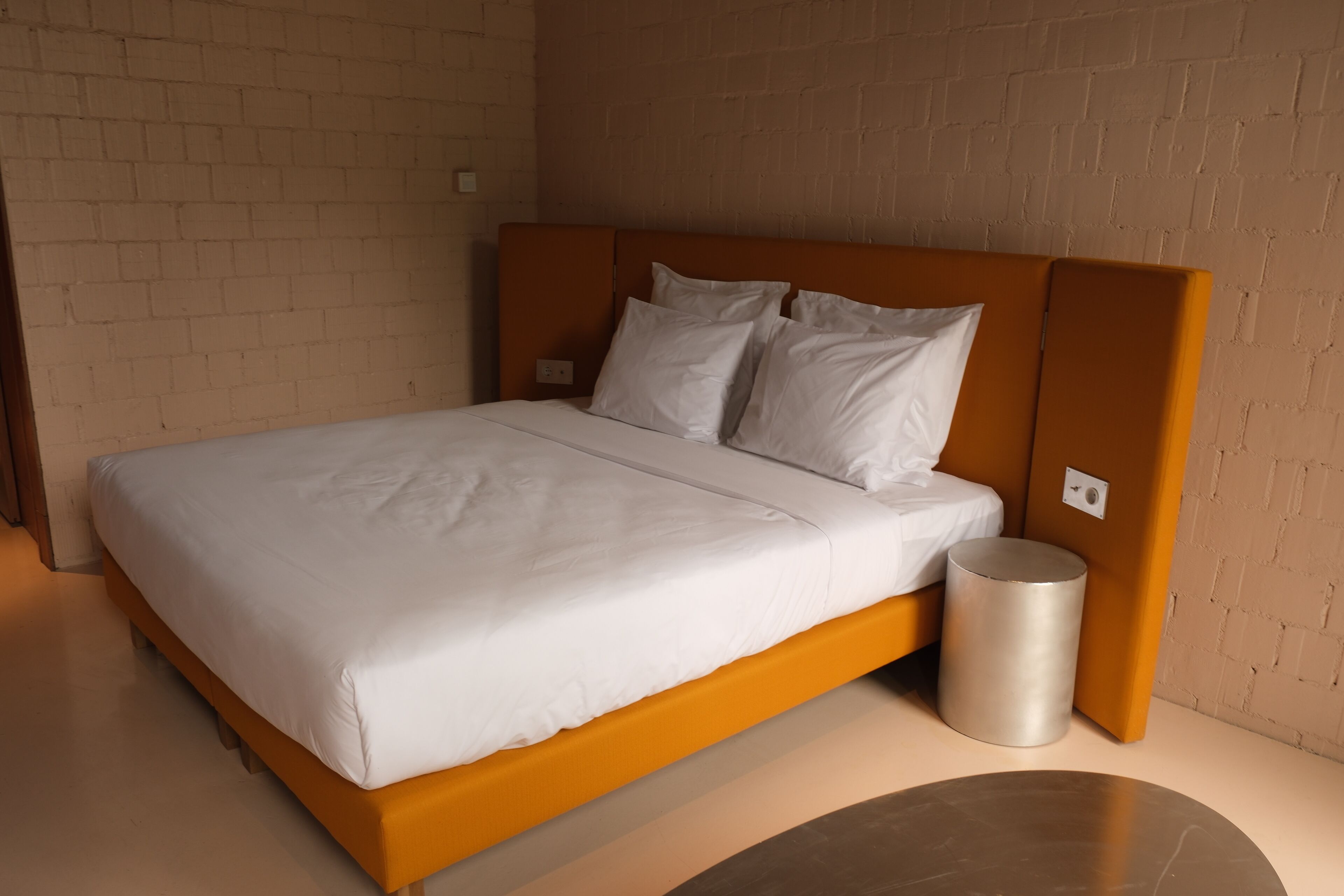 A modern bedroom with a brick wall, a wooden headboard, a white bedspread, and a metallic trash bin.