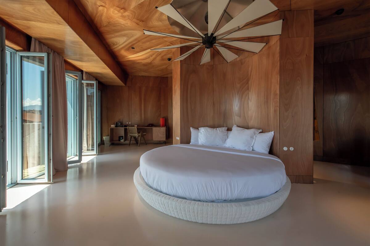 Un dormitorio moderno con una cama redonda grande, techo de madera y ventanas amplias.