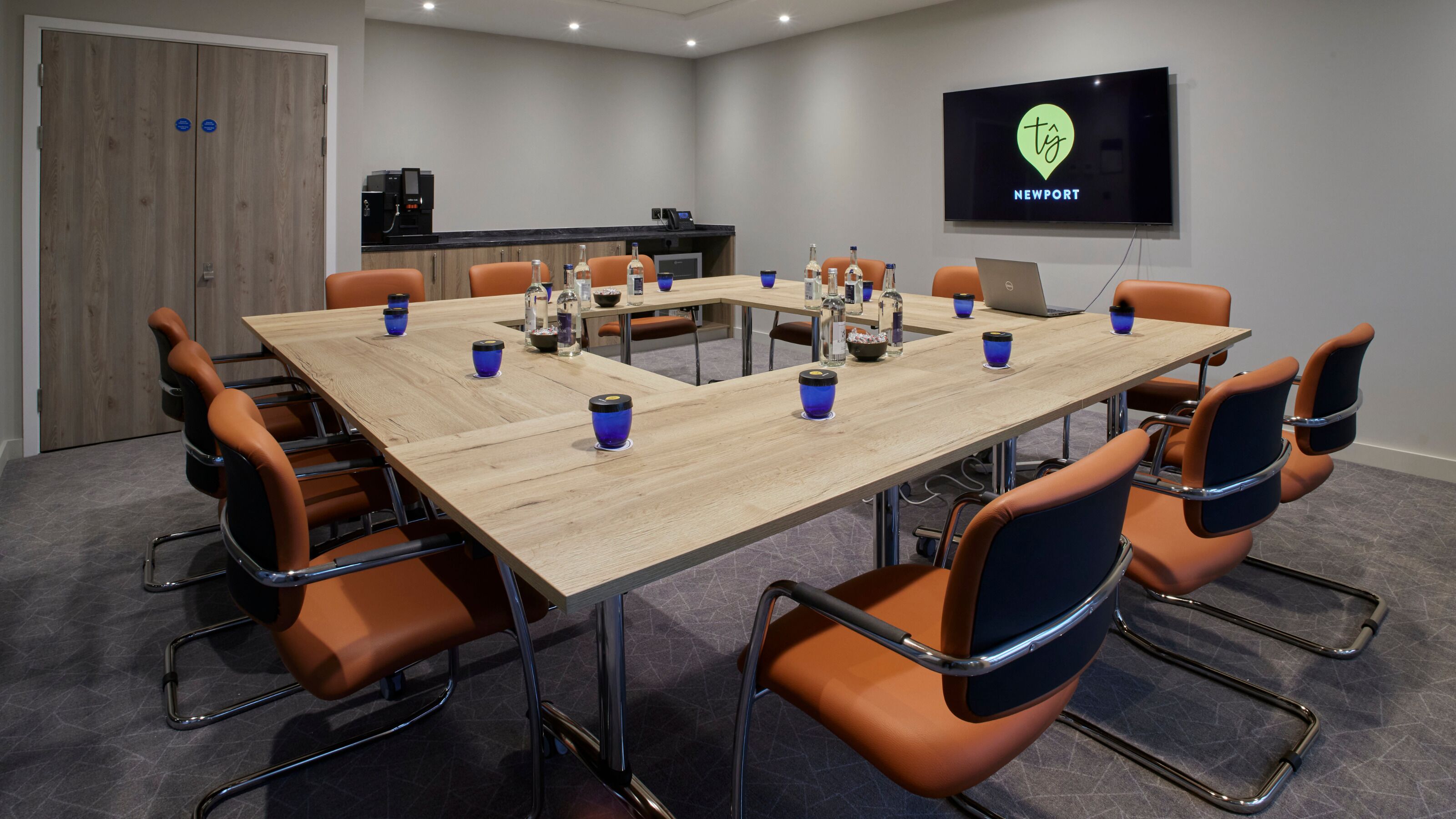 A conference room with a long table, orange chairs, a laptop, and a TV displaying 'NEWPORT'.