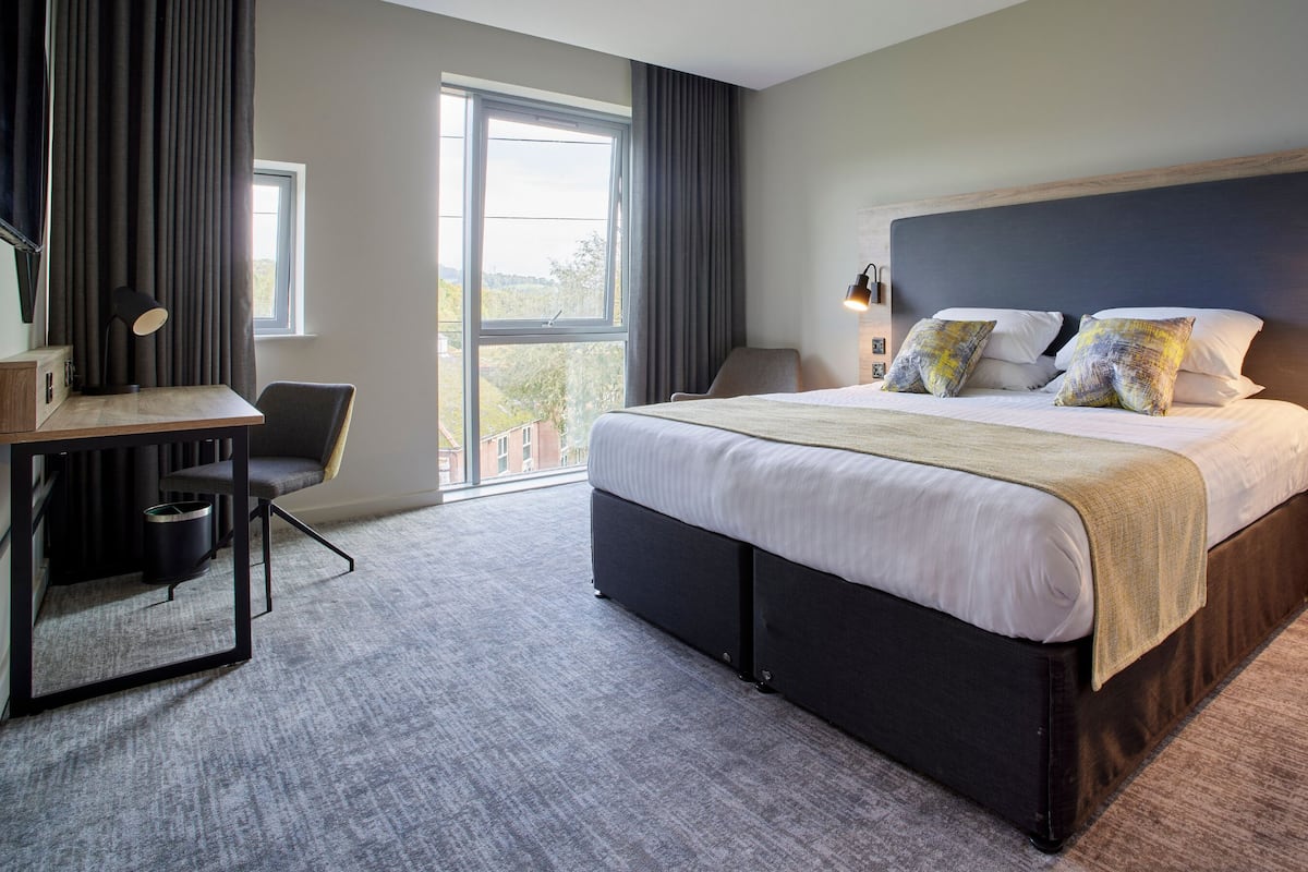 A modern hotel room with a large bed, a desk, and a chair. There is a window with curtains, a nightstand with a lamp, and a small trash can.