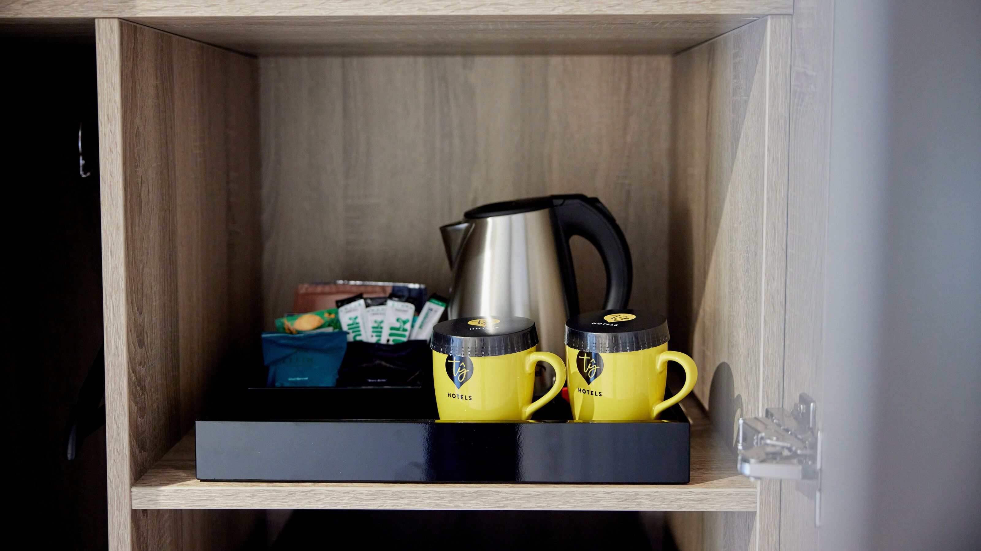 A wooden cabinet with a black tray containing two yellow mugs, a silver kettle, and some packaged food items.