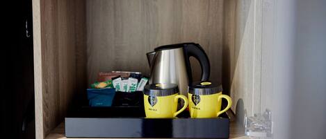 Un mueble de madera con una bandeja negra que contiene dos tazas amarillas, un hervidor plateado y algunos alimentos envasados.