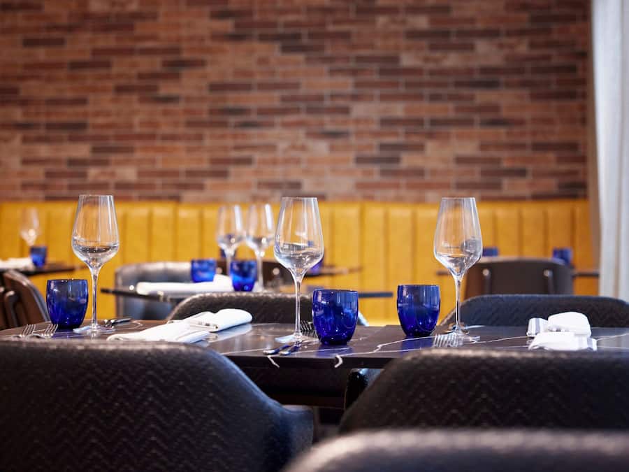 A restaurant with tables set for dining, featuring a brick wall background and blue glassware.